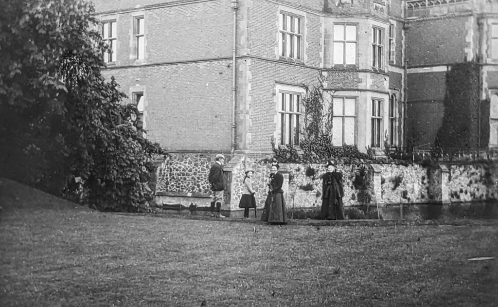An Edwardian photo of a house with moat in black and white. There are four people standing on the lawn.